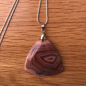 Op de afbeelding: Een zilveren ketting met een hanger gemaakt van een grote, bruine, witte en oranje agaat steen. De steen heeft een uniek, gelaagd patroon.