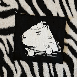 May include: Black square patch with a white illustration of a capybara and oranges. The capybara is in water, with oranges floating around it. The patch is set against a zebra-striped background.
