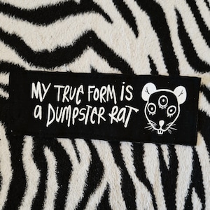 May include: Black and white iron-on patch with the text "My true form is a dumpster rat" and a white cartoon rat with three eyes.