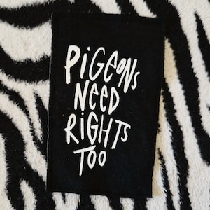 May include: A black rectangular patch with the white text "Pigeons Need Rights Too." The patch is placed on a black and white zebra-striped background.