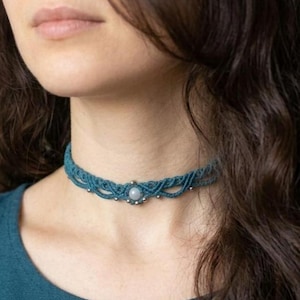 Può includere: Una collana girocollo in macramè color teal con una pietra centrale rotonda e chiara. La collana è indossata intorno al collo. Il design in macramè presenta piccole perline color argento. Lo sfondo è sfocato.