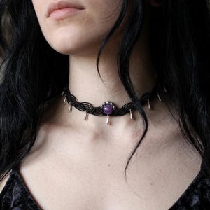 Può includere: Girocollo nero con una pietra centrale viola e pendenti a goccia color argento. La collana è realizzata in cordoncino nero intrecciato. Il gioiello è in stile gotico.