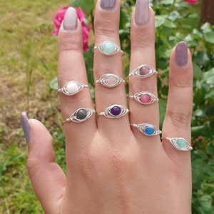 Può includere: Una mano indossa otto anelli d'argento con pietre preziose di diversi colori. Gli anelli sono tutti avvolti in filo e presentano una singola pietra preziosa al centro di ogni anello. Le pietre preziose sono verdi, rosa, bianche, grigie, viola, blu e rosse.