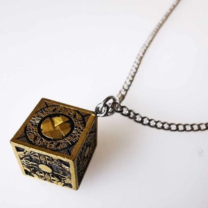 Puede incluir: Un collar dorado con un colgante en forma de cubo que presenta detalles intrincados y un diseño circular en el centro. El colgante está unido a una cadena plateada.