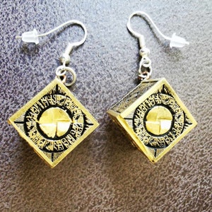 May include: A pair of gold-colored, square earrings with black etched designs. Each earring features a central circular design with a cross-like pattern and is suspended from a silver-colored hook.