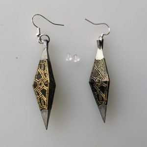 May include: A pair of black and gold earrings with a geometric design. The earrings are shaped like a diamond and have a pointed tip. The earrings are hanging from silver hooks.