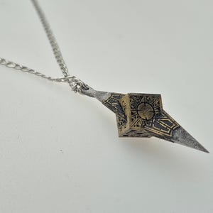 Puede incluir: Un collar plateado con un colgante geométrico. El colgante presenta un diseño de cubo con patrones detallados y un motivo circular central. El colgante está unido a una delicada cadena, creando un accesorio llamativo.