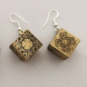 May include: A pair of gold-colored cube earrings with intricate black etched designs. The earrings have silver-colored ear wires. The cubes feature detailed patterns on each face, creating a unique and eye-catching accessory.