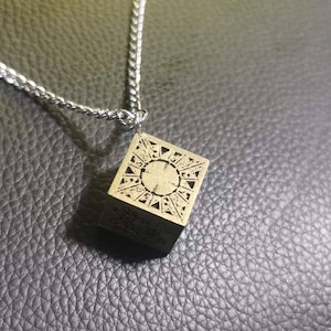 Puede incluir: Un collar de cadena plateada con un colgante dorado en forma de cubo con diseños intrincados.