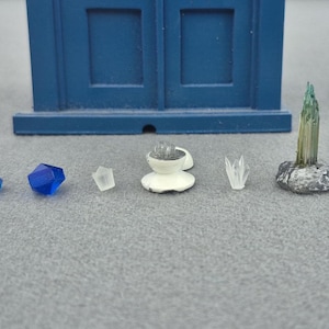 May include: A selection of miniature objects, including crystals, a teacup, and a small, blue door. The objects are arranged on a grey surface. The objects include a yellow and black striped bead, a clear plastic structure, and a blue crystal.