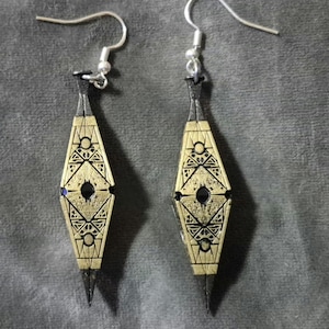 May include: A pair of gold and black geometric earrings with a silver hook. The earrings are shaped like a diamond with intricate patterns.