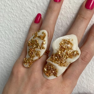 Può includere: Due anelli in resina bianca dalle forme irregolari, decorati con dettagli in foglia d'oro. Gli anelli sono mostrati su una mano con smalto rosa acceso. Un gioiello unico.