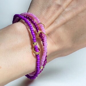 May include: A set of three beaded bracelets in shades of pink and purple with gold accents. The bracelets are stacked on a person's wrist.