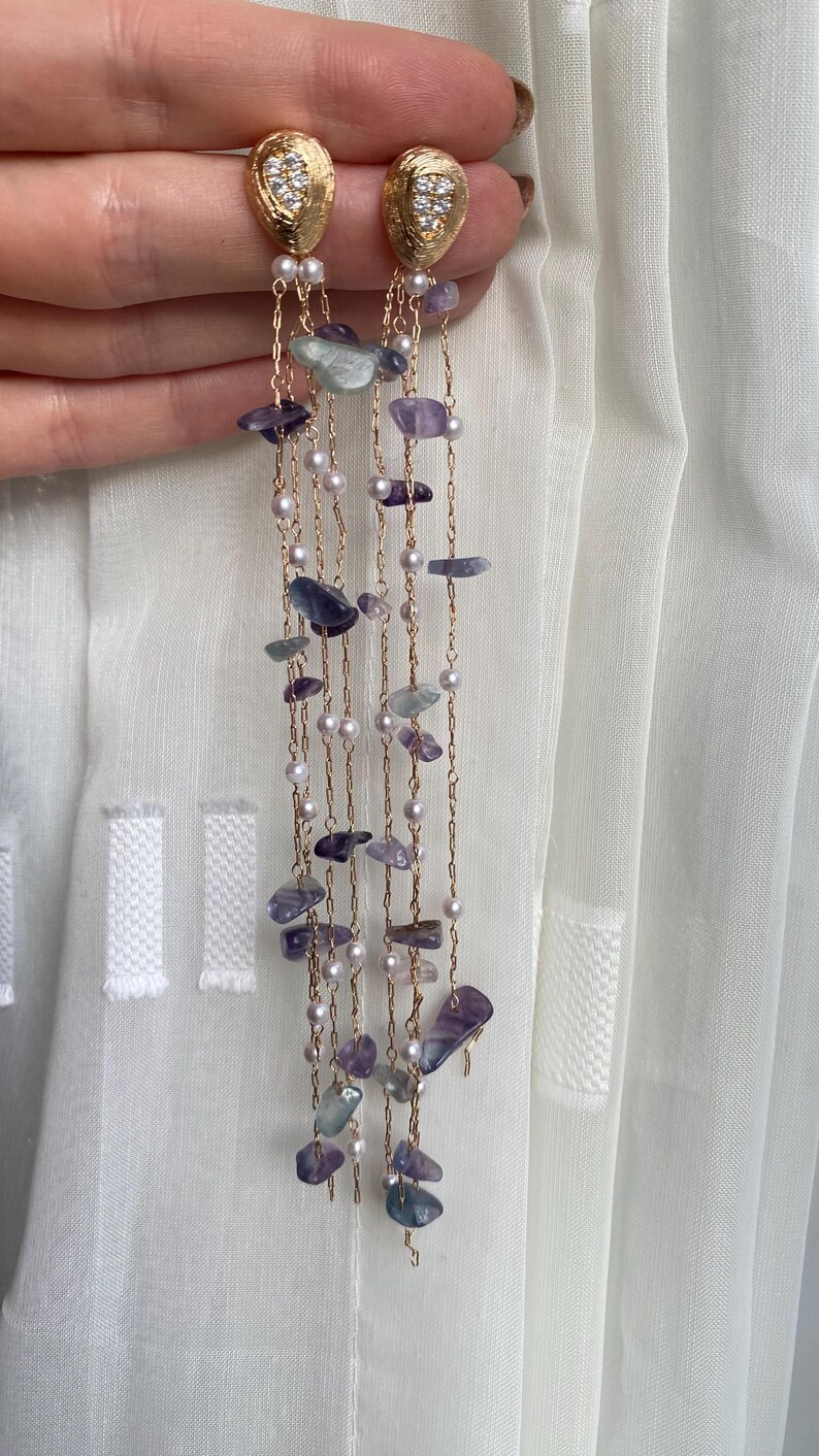 May include: Gold-toned earrings with a textured oval top, adorned with small clear stones. Delicate chains cascade down, embellished with small pearls and irregularly shaped purple and green stones. The earrings are displayed against a white background.