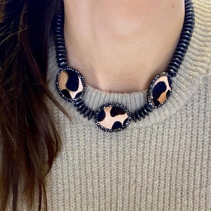 Peut inclure: Un collier de perles noires avec trois pendentifs ronds ornés d'un motif léopard. Le collier est porté par une personne aux cheveux longs et bruns.