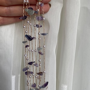 May include: Gold-toned dangle earrings with a textured oval top, adorned with small clear stones. The earrings feature strands of small pearls and irregularly shaped purple and blue stones, creating a cascading effect. The earrings are held against a white background.
