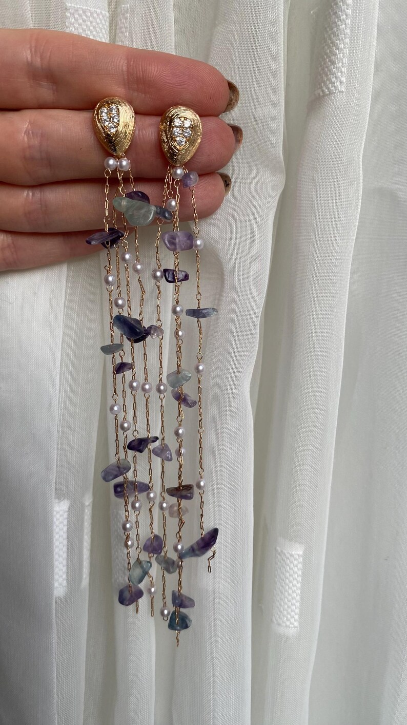 May include: Gold-toned dangle earrings with a textured oval top, adorned with small clear stones. The earrings feature strands of small pearls and irregularly shaped purple and blue stones, creating a cascading effect. The earrings are held against a white background.