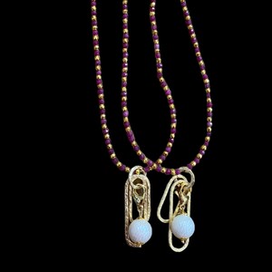 May include: Two gold necklaces with purple beads and a white pearl pendant on each. The necklaces are hanging against a black background.
