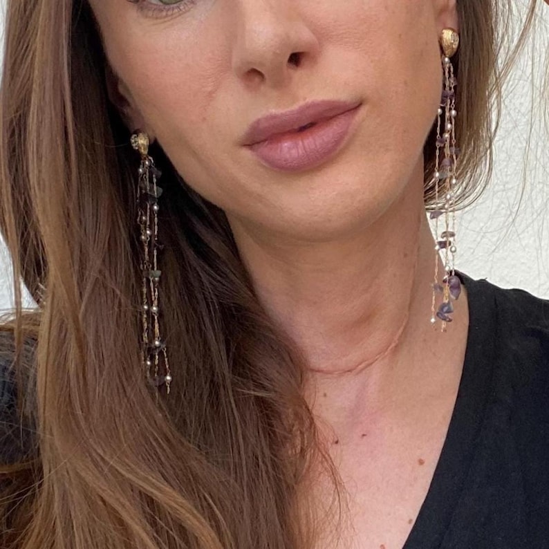 May include: Gold-toned earrings with multiple strands of beads and small stones. The earrings have a textured, round stud and dangle down, showcasing a mix of colors including purple, green, and silver. The earrings are a statement piece.