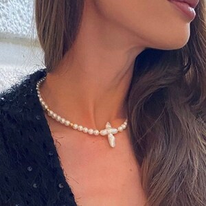 Op de afbeelding: Een parelketting met een grote, onregelmatige parel kruis hanger. De ketting is gemaakt van kleine, witte parels en gouden kralen.