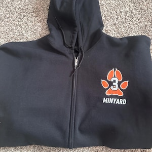 Puede incluir: Sudadera con capucha negra con cremallera y capucha con cordón. La sudadera presenta un diseño de huella de pata en naranja y blanco con el número 3 en el interior. La palabra "MINYARD" está impresa debajo.