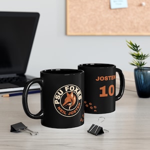 Puede incluir: Dos tazas de cerámica negras. Una taza presenta un logotipo negro y naranja con el texto "PSU Foxes Neil Josten" y huellas de patas. La otra taza tiene el texto "Josten 10" en naranja.