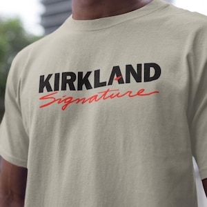 May include: A beige t-shirt with the words "KIRKLAND Signature" printed on the front in black and red. The shirt is short-sleeved and appears to be made of a soft material. The shirt is being worn.
