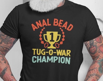 May include: Black t-shirt with the text "ANAL BEAD TUG-O-WAR CHAMPION" in orange, yellow, and teal. The design includes a trophy and a beaded circle around the words "ANAL BEAD".