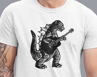 May include: A light gray t-shirt featuring a black illustration of Godzilla playing a guitar. The design is centered on the shirt. The person in the image is wearing blue jeans and has a tattoo on their left arm.