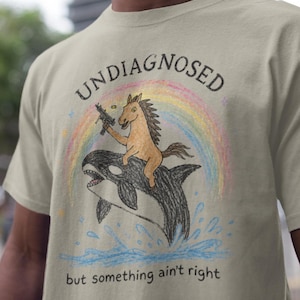 Può includere: Una t-shirt beige con un disegno a pastello di un cavallo marrone che cavalca un'orca sotto un arcobaleno. Il cavallo tiene in mano un'arma da fuoco. Il testo recita UNDIAGNOSED in alto e but something ain't right in basso in nero.