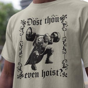 May include: A light beige t-shirt with a black illustration of a knight performing a barbell squat. The text "Dost thou even hoist?" is printed above and below the knight, framed by decorative flourishes.