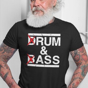 Puede incluir: Camiseta negra con la frase "DRUM & BASS" en blanco, con las palabras tachadas en rojo. La persona que lleva la camiseta tiene barba blanca y tatuajes. La camiseta es una prenda informal.