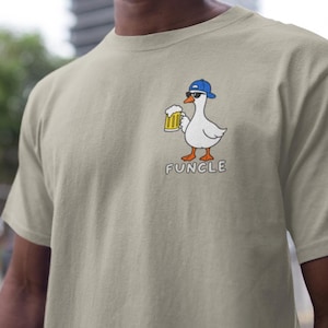 May include: A beige short-sleeved t-shirt featuring a graphic of a white goose wearing a blue baseball cap and black sunglasses while holding a mug of beer. Below the goose, the word "FUNCLE" is printed in white capital letters.