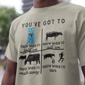 Op de afbeelding: Lichtbeige t-shirt met de tekst "YOU'VE GOT TO" boven vier panelen. Elk paneel heeft een koe-illustratie en de tekst: "Know when to hold 'em", "Know when to cull 'em", "Know when to walk away" en "Know when to run".