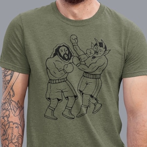 Könnte beinhalten: Ein grünes T-Shirt mit einer schwarz-weißen Illustration von zwei Boxern, die kämpfen. Der eine Boxer hat einen Bart und der andere Hörner.