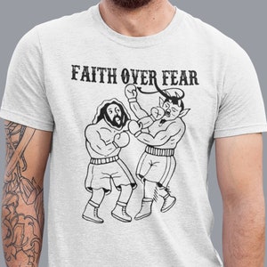 May include: A white t-shirt featuring a black and white illustration of two boxers. The text "FAITH OVER FEAR" is printed above the illustration.