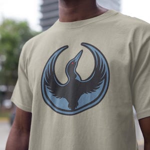 May include: A beige t-shirt featuring a stylized bird emblem in black, light blue, and red. The bird is depicted with outstretched wings within a light blue circle. The t-shirt is made of a soft material.