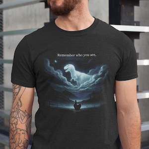 Puede incluir: Camiseta gris oscuro con una ilustración surrealista. Un Tyrannosaurus Rex fantasmal emerge de las nubes sobre un pollo, con el texto "Remember who you are." El diseño es en tonos azules y blancos.