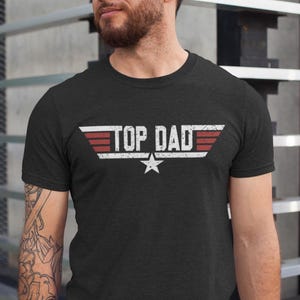 May include: Dark gray t-shirt with the text "TOP DAD" in white, red, and gray, featuring a star graphic. The design is reminiscent of a classic aviation style. A casual, everyday wear tee.