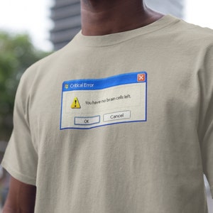 以下が含まれることがあります： ベージュのTシャツ。青と白の「Critical Error」ポップアップウィンドウのグラフィックが特徴です。ウィンドウには黄色の警告サインと「You have no brain cells left.」というテキストが表示されています。