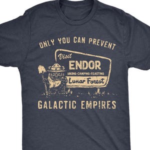 Puede incluir: Camiseta azul marino con un diseño gráfico de estilo vintage. El diseño presenta el texto "ONLY YOU CAN PREVENT" encima de un letrero que dice "Visit Endor, Hiking-Camping-Feasting, Lunar Forest". Debajo del letrero, dice "GALACTIC EMPIRES".