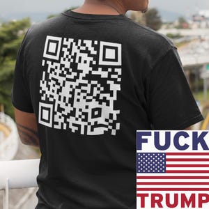 Puede incluir: Camiseta negra con un gran código QR blanco en la espalda. La camiseta también presenta el texto "FUCK TRUMP" con un diseño de la bandera estadounidense. La persona lleva gafas y un pendiente.