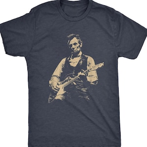 Peut inclure: T-shirt bleu marine avec une illustration s&eacute;pia d'une personne jouant de la guitare &eacute;lectrique. L'illustration repr&eacute;sente une personne en gilet et chemise, tenant la guitare. Le t-shirt est &agrave; col rond.
