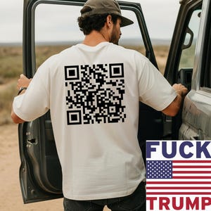 May include: Cream-colored t-shirt with a large black QR code on the back. The shirt also features a graphic with the text "FUCK TRUMP" and an American flag design. The person is standing next to a vehicle.