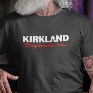 May include: Black t-shirt with the text "KIRKLAND Signature" in white and red. The man is smiling and leaning on a bar. The man has a long white beard and tattoos on his arms.