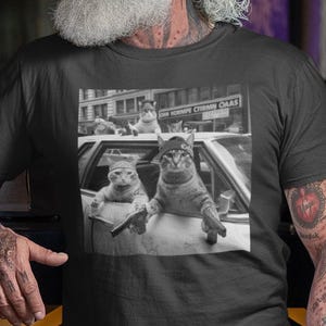 May include: A black t-shirt featuring a black and white image of cats in a car. The image shows two cats in the front seat and two cats on the roof of the car. The person wearing the shirt has tattoos on their arms.