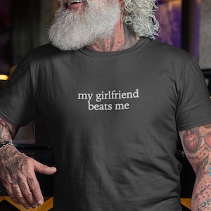 以下が含まれることがあります： 「my girlfriend beats me」の白文字がプリントされた濃いグレーのTシャツ。長い白ひげと腕にタトゥーのある人が着ています。