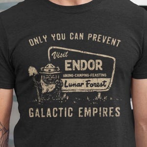 Puede incluir: Camiseta gris oscuro con un diseño gráfico de estilo vintage. El diseño presenta el texto "ONLY YOU CAN PREVENT" encima de un letrero que dice "Visit ENDOR Lunar Forest". Debajo del letrero está el texto "GALACTIC EMPIRES."