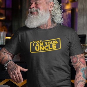 May include: Dark gray t-shirt with the text "LUKE, I AM YOUR UNCLE" in yellow, inside a yellow rectangle. The person wearing the shirt has a long white beard and tattoos on their arm.