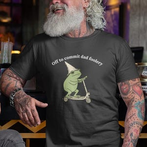 Peut inclure: T-shirt noir avec l'inscription "Off to commit dad foolery" et une grenouille de dessin animé sur un scooter. La grenouille porte un chapeau de sorcier et des lunettes de soleil. Le t-shirt est en tissu doux.